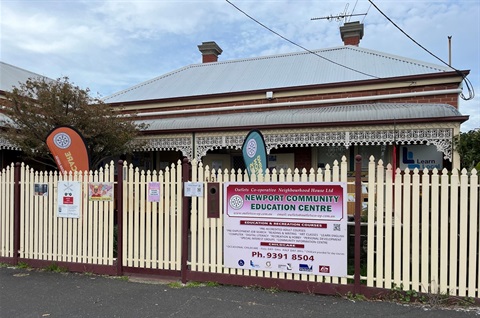 Front view of the Outlets Co-operative Neighbourhood House in Newport, Melbourne. The image shows a heritage-style red brick building with decorative white lacework verandah, cream picket fence and community signage.