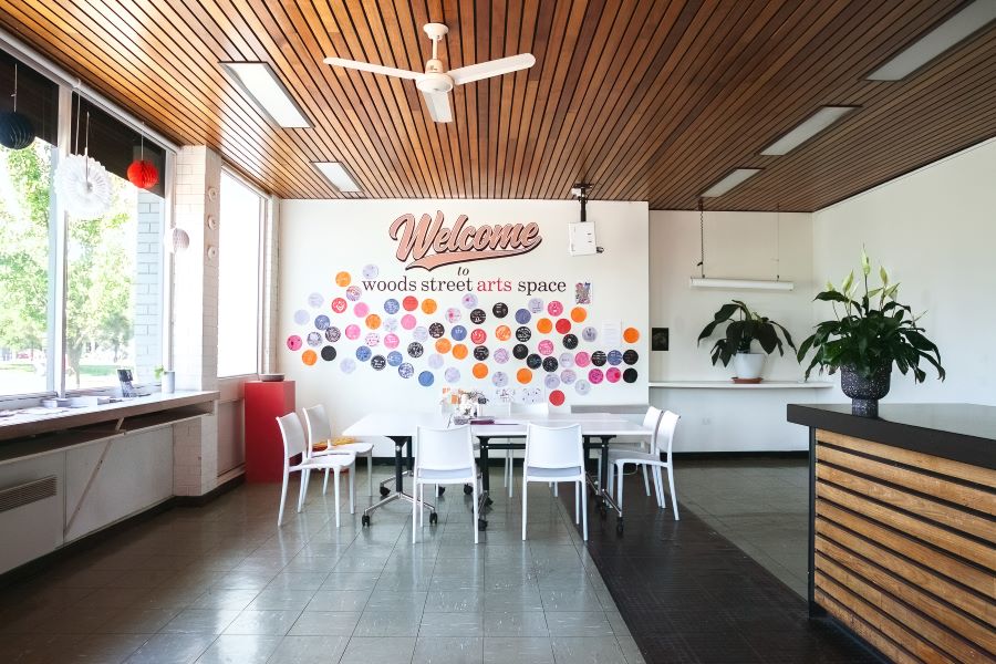 An interior view of the Woods Street Arts Space foyer, with a communal table and chairs, a timber slatted ceiling and grey laminex flooring. A hand-painted sign on the wall reads 